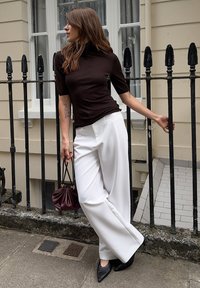 Brązowa koszulka z krótkim rękawem, golf, białe spodnie typu wide-leg, czarne szpilki z ostrym noskiem oraz bordowa torebka, na tle balustrad.