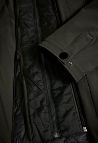 Chaqueta aislante verde oscuro con una textura suave, que presenta cremalleras negras, un puño doblado con un botón a presión y un forro acolchado.