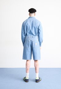 Lyseblå denimskjorte med knaplukning ved håndleddene, parret med matchende denimshorts. Sorte og grønne sneakers, hvide ankelstrømper og en ensfarvet baggrund.