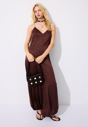 Femme blonde portant une combinaison ample sans manches de couleur marron, un collier ras du cou noir, un collier, tenant un sac à main noir, debout contre un mur blanc.