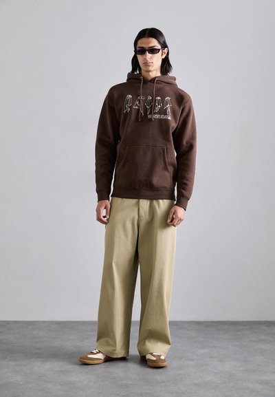 Brown hoodie with white graphic print, kangaroo pocket, beige wide-leg pants, and brown slip-ons with white accents. Smooth texture.