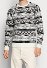 Neulottu neule, jossa on musta, ruskea ja valkoinen geometrinen kuvio; pyöreä kaula-aukko; neulotut resorireunukset hihansuissa ja helmassa; pehmeä tekstuuri.