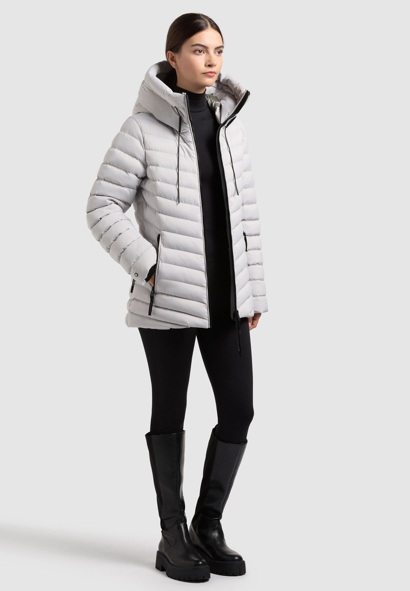 Giacca piumino grigia chiara con cappuccio, design trapuntato orizzontale, chiusura con cerniera e tasche laterali. Abbinata a leggings neri e stivali al ginocchio.