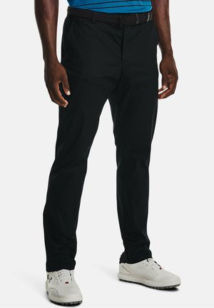 Mann, der schwarze Hose, einen schwarzen Gürtel, weiße Golfschuhe und ein blau gestreiftes Hemd trägt, mit entspannten Händen an den Seiten.