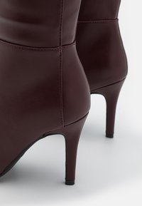 Bottines en cuir bordeaux avec une pointe aiguë, des talons aiguilles hauts, une texture lisse et des détails de couture contrastants.