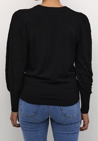 Svart tröja med texturerade, puffiga ärmar. Fitted design, ribbad halsringning och fåll. Buren med blå jeans mot en neutral bakgrund.