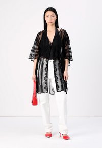 Svart genomskinlig kimono med intrikata spetsdetaljer och batwing ärmar, bärs över en svart topp och matchas med vita byxor och röda accenter.