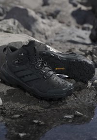 Zwarte trailrunning schoenen met robuuste zolen die rusten op nat rotsachtig terrein bij een kleine plas.