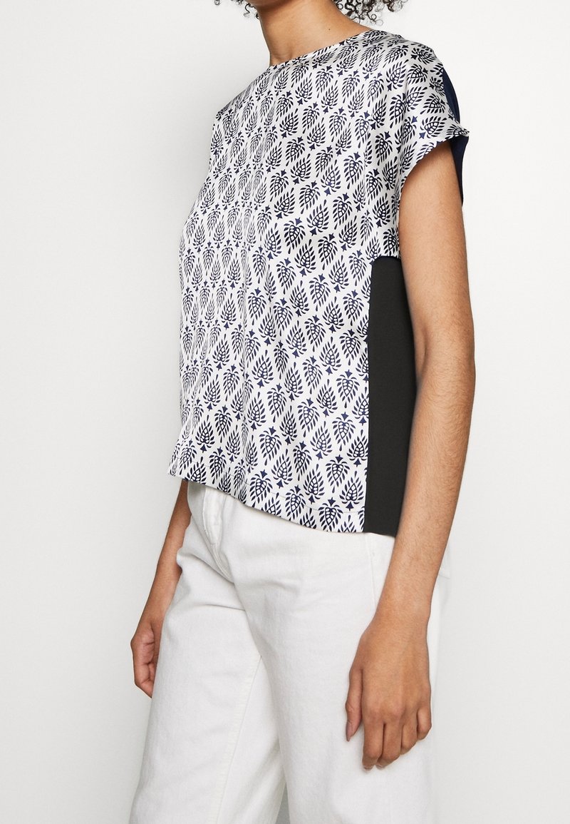 Femme portant un chemisier blanc à manches courtes avec des motifs de feuilles noirs et des panneaux latéraux noirs, associé à un pantalon blanc, sur un fond uni.