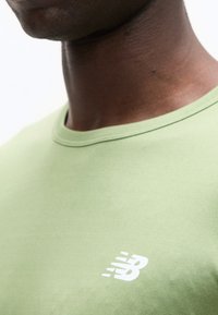 Κοντινό πλάνο του λαιμού και του άνω μέρους του στήθους που φοράει ανοιχτοπράσινο μπλουζάκι New Balance με λευκό λογότυπο στο στήθος.