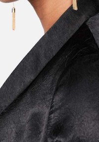 Blazer nero caratterizzato da una texture satinata e colletto a falde. Orecchini con nappine bronzo aggiungono accenti metallici al look.