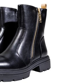 Par de botas negras de cuero hasta el tobillo con suelas de goma gruesas y cremalleras doradas en los laterales sobre un fondo blanco.