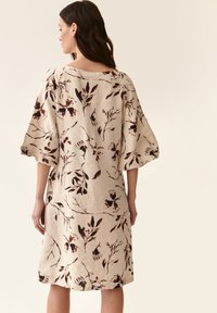 Vaalean beige mekko löysällä leikkauksella, jossa on tummia kukkakuoseja, leveät hihat ja pyöreä pääntie. Valmistettu hengittävästä kankaasta.