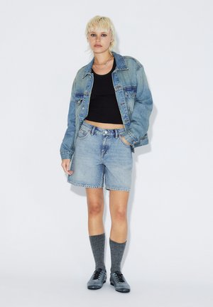 Person iført lyseblå denimjakke og shorts, sort crop top, grå knæstrømper og grå sneakers står mod hvid baggrund.