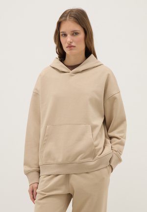Jeune femme portant un sweat à capuche beige et un pantalon assorti, debout avec une expression neutre et une main dans la poche, sur un fond uni.