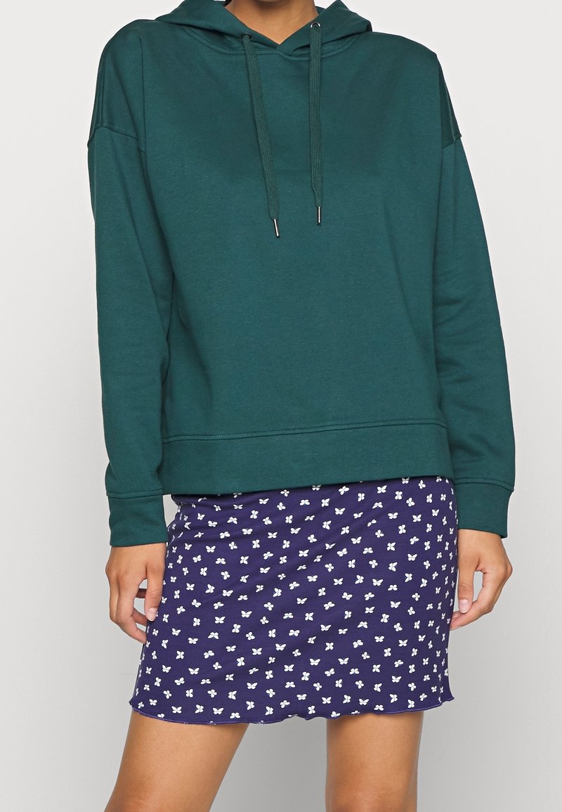 Mörkgrön hoodie med dragkedja och avslappnad passform, i kombination med en marinblå kjol med ett vitt fjärilsmönster.