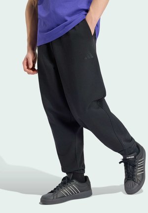 Osoba nosząca czarne spodnie Adidas z logo i czarne buty Adidas z białymi paskami, stojąca z jedną ręką w kieszeni.