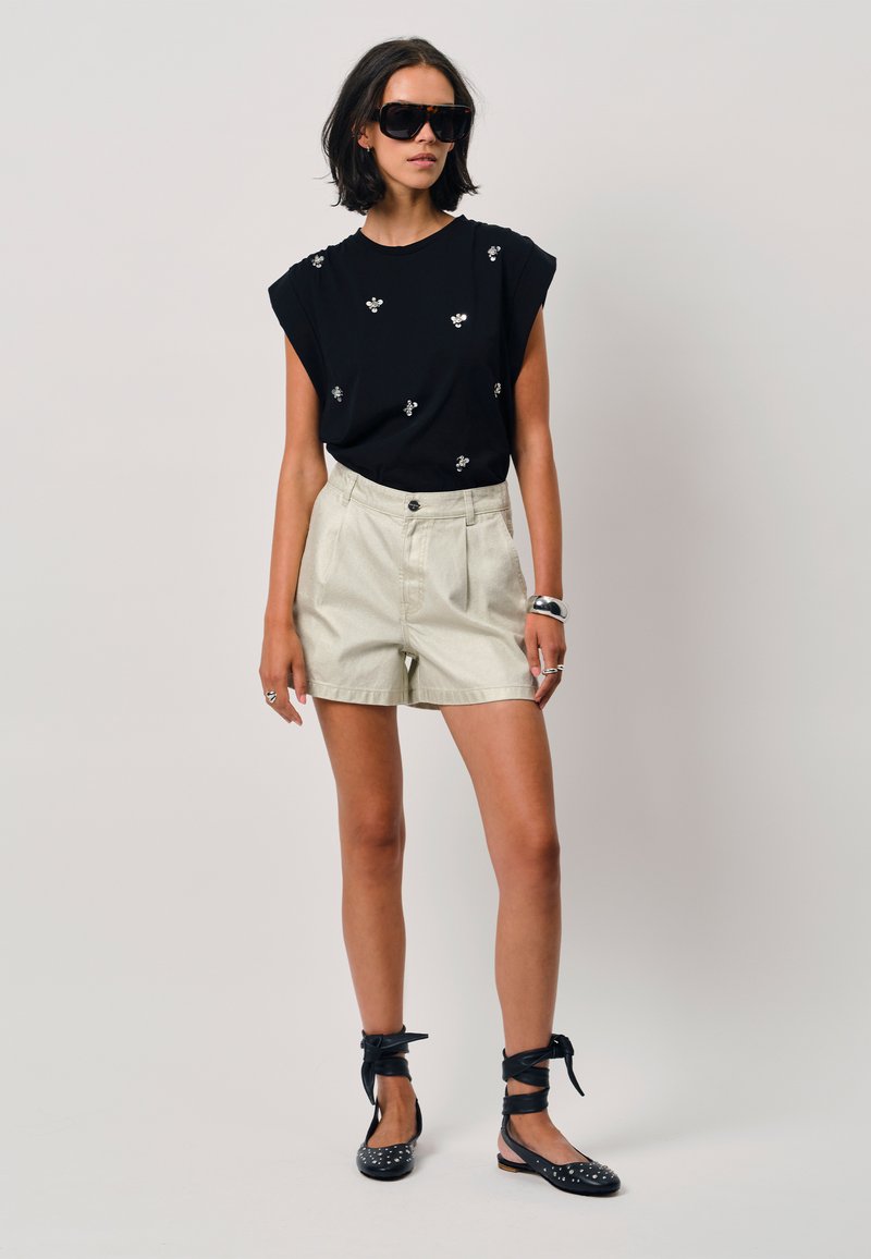 Zwarte top met korte mouwen en bloemenversieringen, gecombineerd met lichtbeige high-waisted shorts. Zwarte instappers met bandjes en studs.