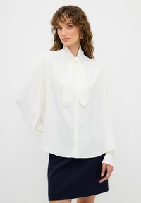 Blouse blanche avec un col à nœud, manches bouffantes longues et coupe ample, associée à une jupe bleu foncé. Tissu lisse avec un léger éclat.