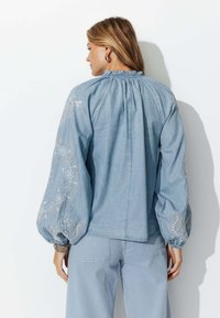 Blouse bleu clair avec un col à volants et des détails brodés sur les manches. Coupe ample et tissu texturé.