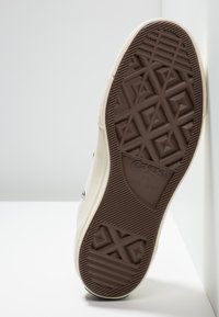 Suola in gomma marrone con motivi di grip testurizzati, parte superiore in tela beige, occhielli neri circolari e bordi in gomma bianca a contrasto.