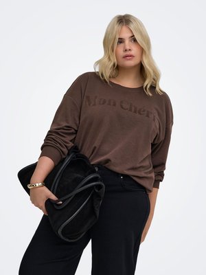 Blonde vrouw met een bruine "Mon Chéri" trui en zwarte broek, die met één arm een grote zwarte tas vasthoudt, staand tegen een witte achtergrond.