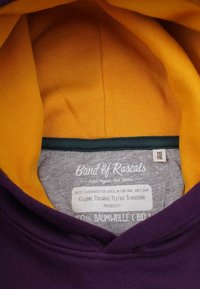 Lila huvtröja med en gul innerfoder. Lappen anger 100% ekologisk bomull, globalt ekologiskt textilstandard och storlek 110/116.