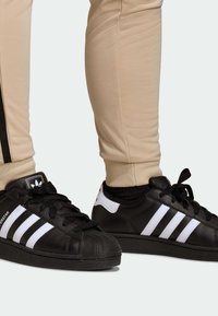 Black leather sneakers feature three white stripes, a textured toe cap, and black laces, paired with beige tapered pants with black accents.