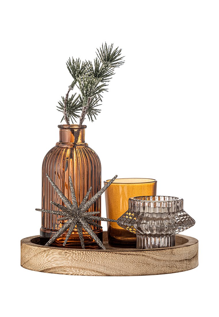 Bottiglie di vetro ambra e portacandele sono disposti su un piatto di legno. Un accento a forma di stella in argento e un rametto di pino completano il set decorativo.