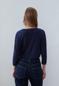 Granatowy sweter w krótkim kroju z długimi rękawami, okrągłym dekoltem oraz teksturowanym splotem. W połączeniu z wysokim stanem ciemnoniebieskich dżinsów z tylnymi kieszeniami.