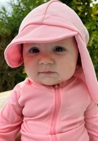 Vaaleanpunainen vetoketjullinen asu, jossa on yhteensopiva leveä lippalakki. Kankaan pinta näyttää teksturoidulta ja kasvoilla on pieni määrä sylkeä.