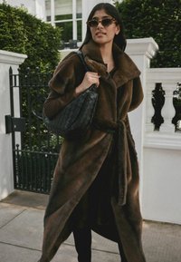 Brown faux fur coat with a wide collar and belt, paired with a woven black handbag and tortoiseshell sunglasses. Outdoor setting.