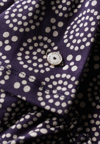 Violetter Stoff mit weißem Punktmuster, versehen mit einem Metallanhänger mit der Aufschrift "LOVE FROM SUPERDRY" und genähten Kanten für Haltbarkeit.