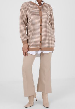 Frau trägt beige Hahnentritt-Cardigan über weißem Hemd, beige weite Hose und beige Stiefeletten mit Absatz vor einfarbigem Hintergrund.