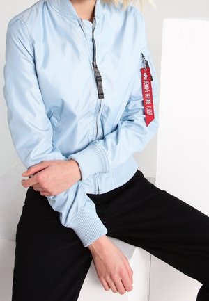 Personne portant une veste bomber bleu clair avec une étiquette rouge "Remove Before Flight" sur la manche, assortie à un pantalon noir, les bras croisés.