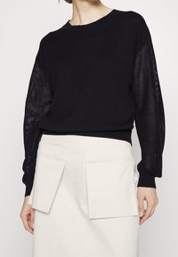 Černý pletený svetr s průhlednými vzorovanými rukávy, kulatým výstřihem a žebrovaným lemem, doplněný světle zbarvenou sukní s dvěma velkými kapsami.