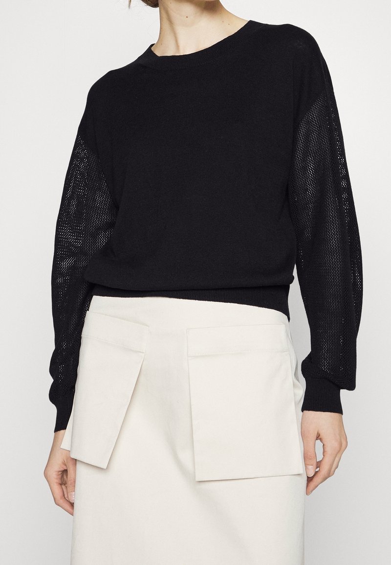 Černý pletený svetr s průhlednými vzorovanými rukávy, kulatým výstřihem a žebrovaným lemem, doplněný světle zbarvenou sukní s dvěma velkými kapsami.