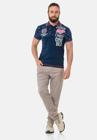 Marineblaues Poloshirt mit kontrastierendem roten und weißen Kragen, versehen mit grafischen Prints und Text, kombiniert mit hellbeigen Hosen und Sneakern.