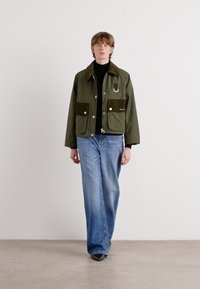 Groene katoenen jas met twee opgestikte zakken, bruine accenten en een zilveren ringdetail, gecombineerd met lichtblauwe wijduitlopende jeans.