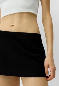 Černá mini sukně s hladkou texturou, bezšvovým designem a přiléhavým siluetou. Žádná viditelná kovová nebo vzorová dekorace.