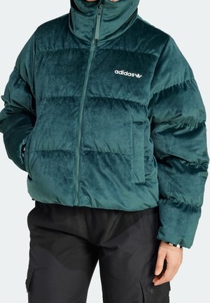 Personne portant une doudoune en peluche Adidas de couleur sarcelle avec un col haut, associée à un pantalon cargo noir, main dans la poche.