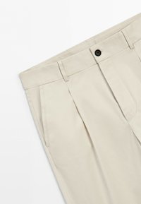 Beige trousers with front pleats, side pockets, belt loops, and a black button closure on a white background.