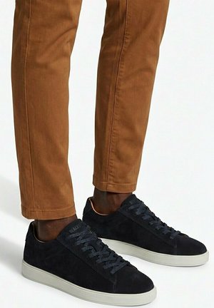 Piernas con pantalones marrones y zapatillas negras bajas con suelas blancas, de pie sobre una superficie gris claro.