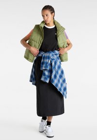 Chaleco de plumas verde sobre una camiseta negra, camisa de cuadros azul atada a la cintura, falda negra larga y zapatillas blancas. Texturas de tela planas y suaves.