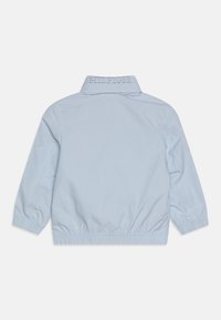 Světlomodrá lehká bunda s stojáčkovým límcem, elastickými manžetami a staženým lemem. Na zadní straně je vyšitý nápis značky.