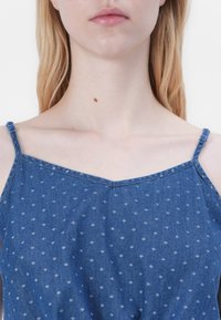 Top din denim cu bretele subțiri, având un decolteu în V și un model alb subtil. Materialul are o textură netedă într-o nuanță de albastru.