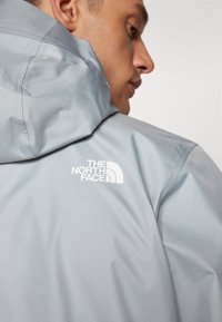 Svetlošedá nepremokavá bunda s kapucňou a bielym logom značky The North Face na ľavom ramene. Hladká textúra, minimalistic design.