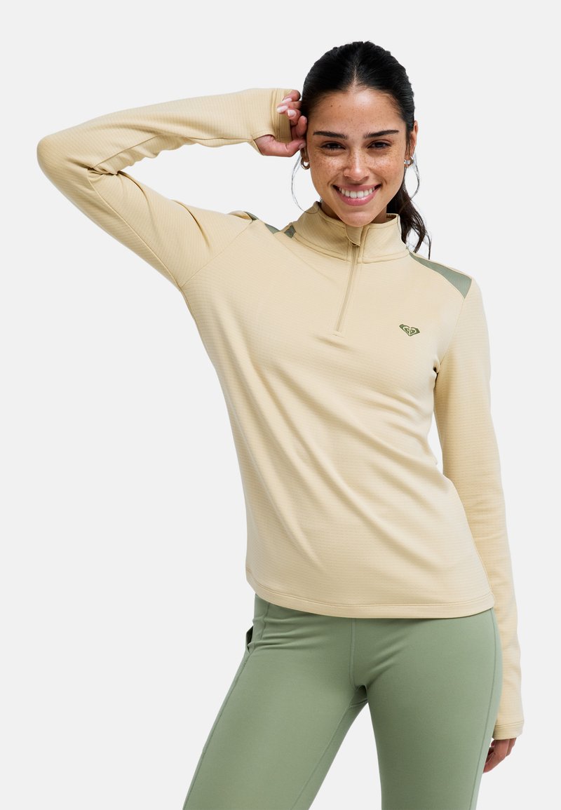 Beiges Langarm-Top mit Viertel-Reißverschluss, gerippter Textur und grünen Akzenten auf den Schultern, kombiniert mit mintgrünen Leggings.