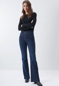 Vaqueros de campana azul oscuro con cintura alta, costuras en contraste y estilo de cinco bolsillos, combinados con una blusa negra de manga larga y botas negras de tacón.