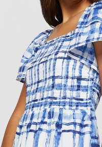 Robe à carreaux bleu et blanc avec un corsage plissé, des manches tombantes sur les épaules et une jupe évasée. Tissu léger avec un motif texturé.